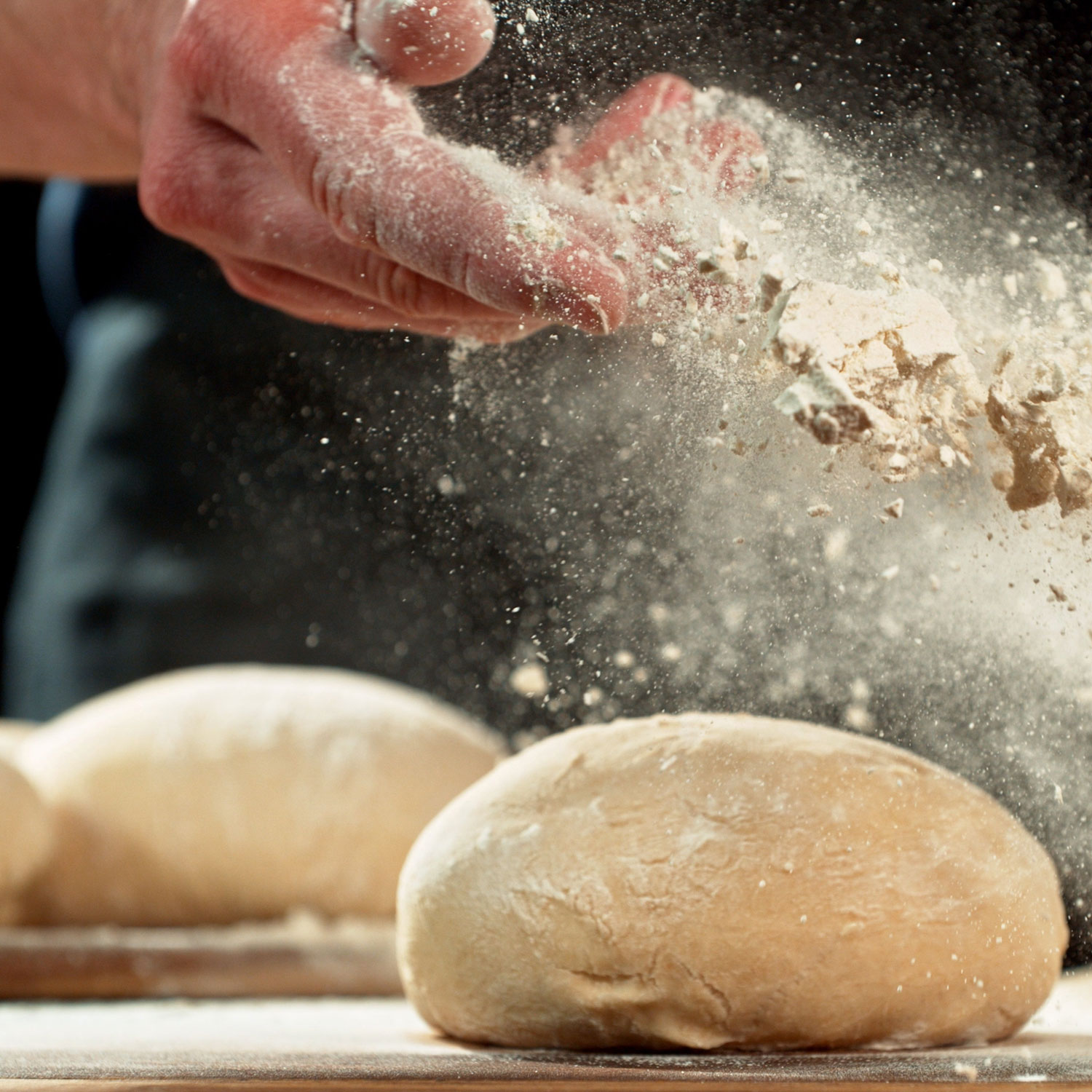 A baker tosses flour over shaped dough loaves, lit warm and low — the moment before the oven.